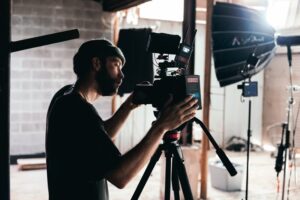 **Alt Text:** A filmmaker with a beard and cap operates a video camera on a tripod in a studio setting.