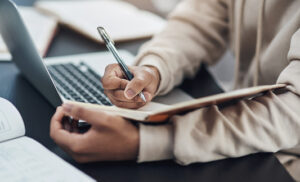 **Alt text:** A person writing in a notebook while using a laptop at a desk.