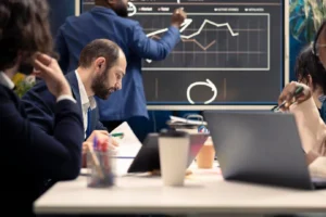 Alt text: A group of business professionals in a meeting with one person pointing at a graph on a digital screen.
