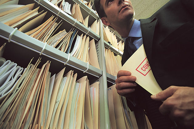 A man in a suit holding a folder labeled "Confidential" stands in front of a shelf filled with numerous file folders.