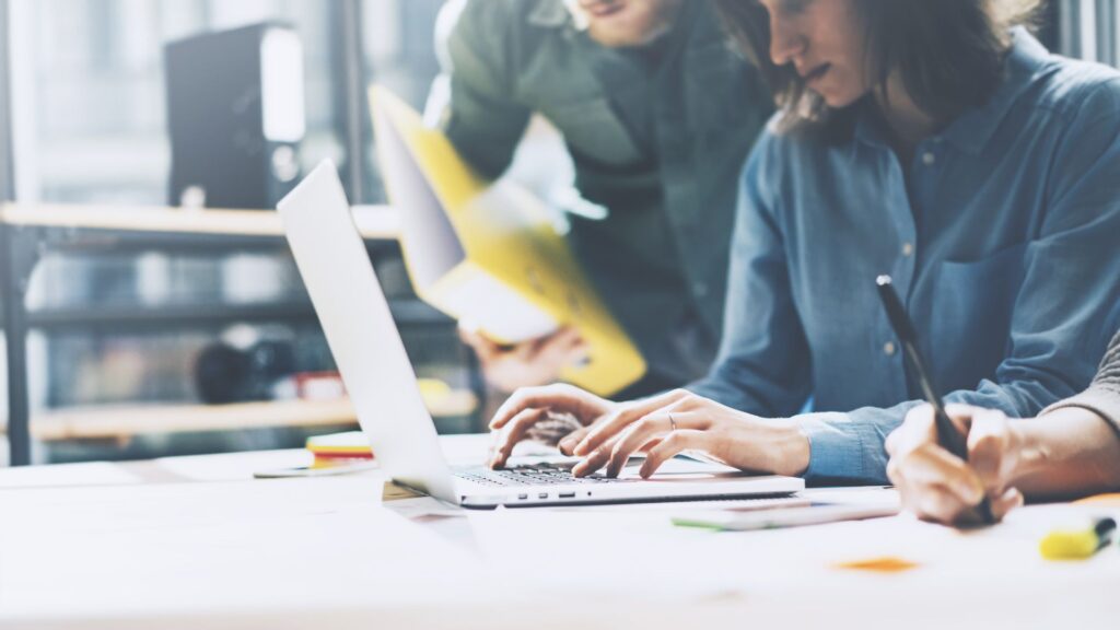 People working together on a project, with one person typing on a laptop and another taking notes.