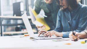 People working together on a project, with one person typing on a laptop and another taking notes.