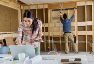 Alt text: A woman looks at a laptop and holds a color palette in a room under construction, while a man works on the wall in the background.