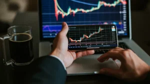 **Alt Text:** A person holds a smartphone displaying a stock market chart in front of a laptop showing a similar chart, with a cup of coffee nearby.