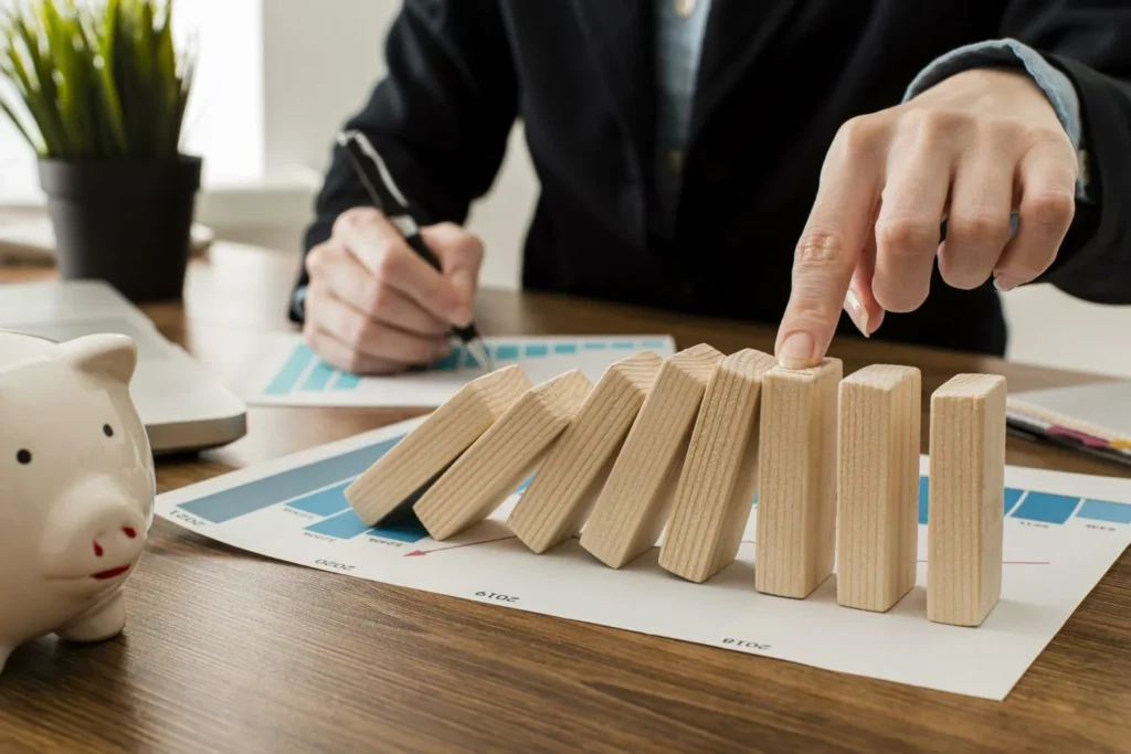 Alt text: A person prevents a series of wooden blocks from toppling over on top of financial charts, with a piggy bank nearby.