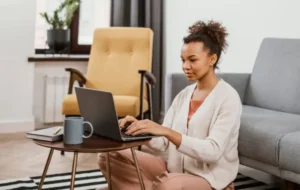 Alt text: A woman sitting on the floor in front of a coffee table, using a laptop in a cozy living room setting.