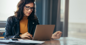 A person with glasses and a leather jacket is working on a laptop at a table.