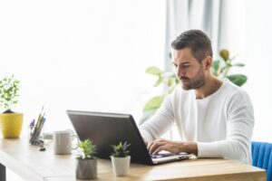 **Alt text:** A man in a white shirt is working on a laptop at a desk adorned with small potted plants and a mug.