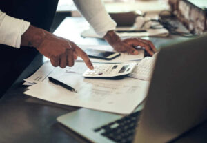 **Alt text:** A person using a calculator while working on financial papers at a desk, with a pen and a smartphone nearby.