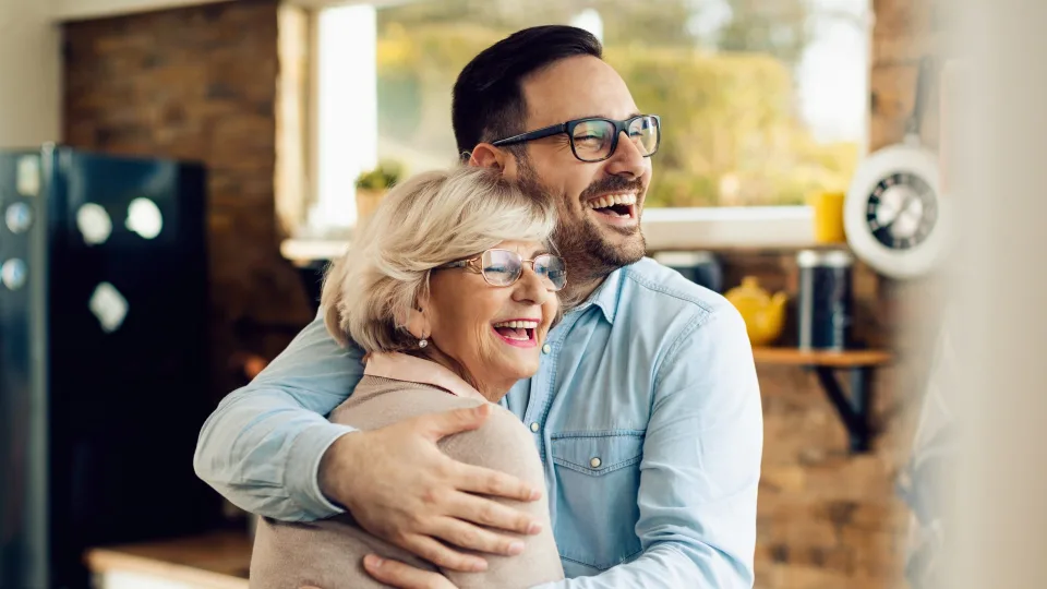 **Alt Text:** A smiling man and an older woman wearing glasses embrace warmly in a cozy kitchen setting.
