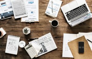 A cluttered wooden desk with a laptop, calculator, newspaper, planner, tax form, coffee cups, smartphone, and various documents.