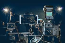 **Alt Text:** A complex camera setup with multiple screens and devices aimed at capturing a scene with two people in the background under bright lights.