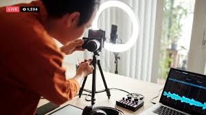 **Alt Text:** A person adjusts a camera on a tripod with a ring light and a laptop displaying audio waveforms in the background, indicating a live streaming setup.