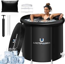 A person is relaxing in a black ArctiPulseX inflatable hot tub, surrounded by accessories such as a pillow, cover, air pump, ice packs, and a repair kit.