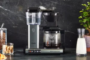 Alt text: A Moccamaster coffee maker is placed on a countertop alongside a jar of coffee beans, a jar of sugar cubes, and a carafe of milk, with a green plant in the background.