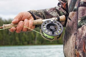 **Alt text:** A person wearing a camouflage jacket holds a fly fishing rod and reel beside a river.