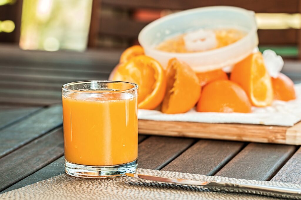 **Alt text:** A glass of freshly squeezed orange juice on a table with halved oranges and a juicer in the background.