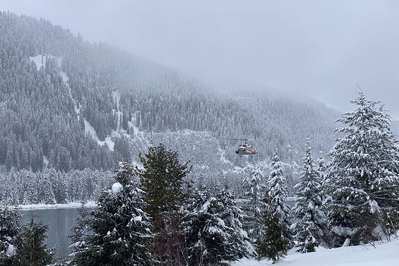 **Alt text:** A helicopter flies over a snowy forest landscape with a misty mountain in the background.