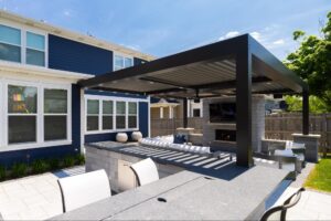A modern outdoor patio with a pergola, seating area, fireplace, and adjacent to a blue house on a sunny day.