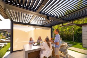 A group of people gather around an outdoor kitchen bar under a modern pergola in a sunny backyard.