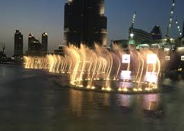 A nighttime view of a brightly lit water fountain display in front of modern skyscrapers and buildings.