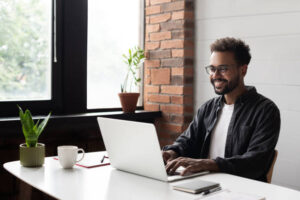A man wearing glasses sits at a desk by a window, working on a laptop with a smile, surrounded by plants, a mug, and a notebook.