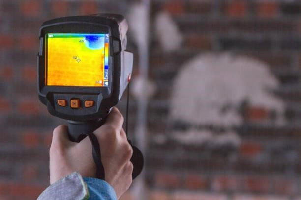 A person holds a thermal imaging camera aimed at a brick wall with the screen displaying a heat map.