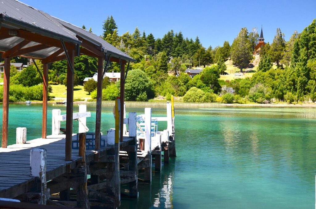 A wooden pier extends over a calm, turquoise lake with lush greenery and a small church visible in the background.