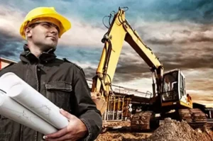 A construction worker in a yellow hard hat holds blueprints while standing in front of an excavator at a construction site.