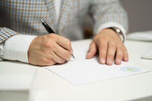A person in a checkered jacket signing a document with a pen on a white desk.