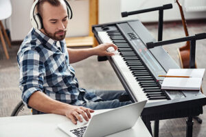 **Alt Text:** A person wearing headphones uses a laptop while sitting at an electronic keyboard with a notebook and pencil on top.