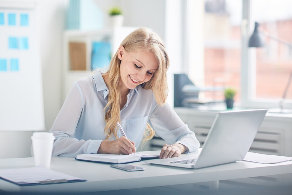 A woman with long blonde hair is sitting at a desk, smiling while writing in a notebook with a laptop and a coffee cup nearby.