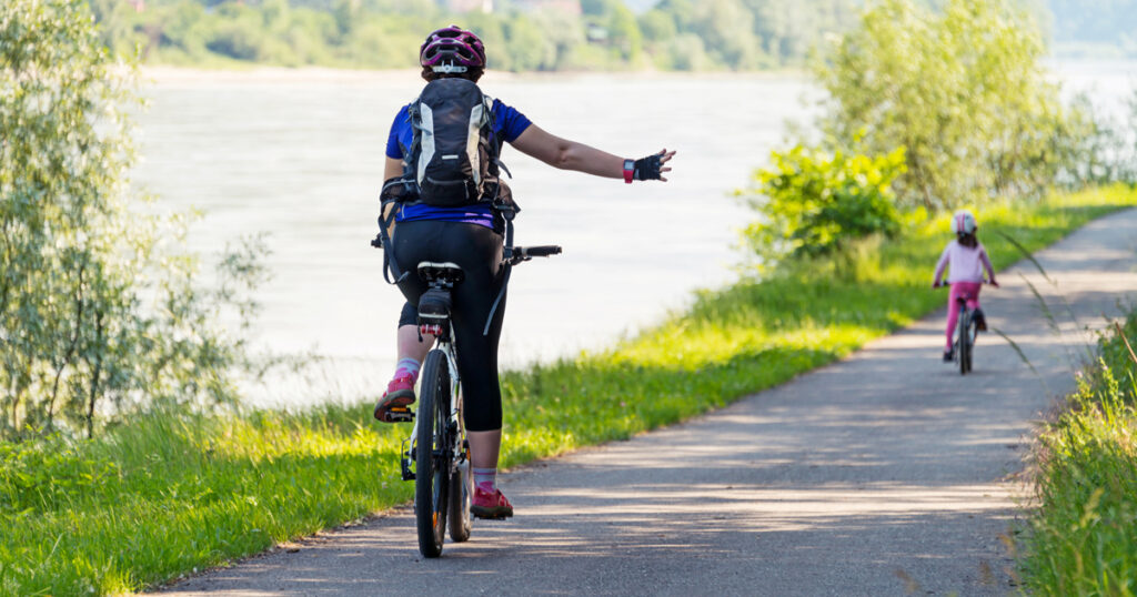 An adult cyclist and a child on bikes ride along a river path surrounded by greenery.