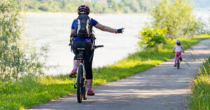 An adult cyclist and a child on bikes ride along a river path surrounded by greenery.