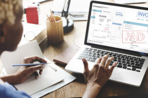 **Alt text**: A person sitting at a desk reviewing a "paid" invoice on a laptop while taking notes.