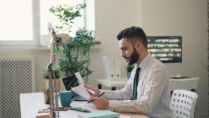 A man in a plaid shirt and tie is sitting at a desk, reviewing papers with a laptop and a plant nearby.