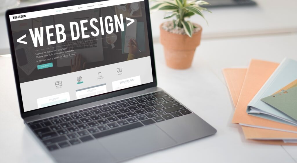 A laptop displaying a web design website sits on a desk alongside a potted plant and a stack of colorful folders.