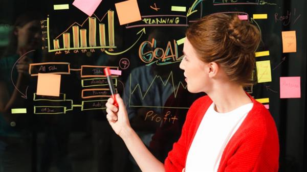 **Alt text:** A woman in a red cardigan writes and points at various colorful sticky notes and diagrams on a glass board in an office setting.