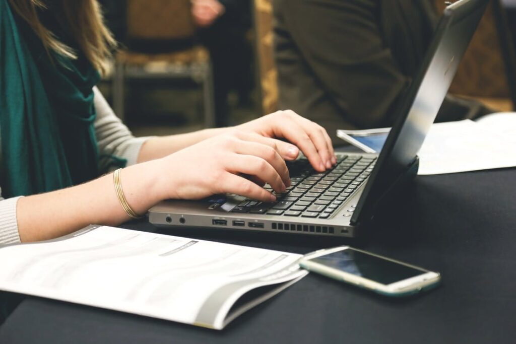 **Alt Text:** A person typing on a laptop with a smartphone and an open book nearby on a table.
