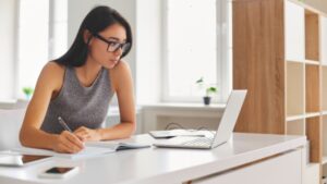 **Alt text:** A woman wearing glasses is working on a laptop at a desk while taking notes in a notebook.
