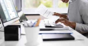 **Alt text:** A person in a suit working at a desk with a computer monitor displaying financial documents, using a calculator and examining papers.