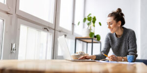 **Alt text:** A woman in a gray sweater is smiling at her laptop while working at a wooden table with a cup and a notebook nearby.