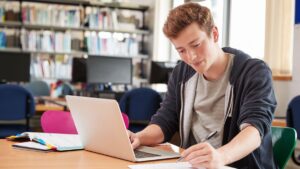 **Alt text:** A young person studies in a library, using a laptop and taking notes.