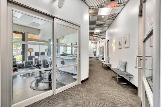 **Alt Text:** A hallway in a modern office building with glass doors to the left revealing an interior office space, and a bench with cushions against the wall on the right.