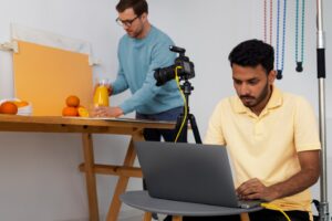 Alt text: A man in a blue sweater arranges oranges and orange juice on a table while another man in a yellow shirt works on a laptop connected to a camera.