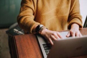 **Alt text**: A person wearing a yellow sweater works on a laptop at a wooden desk.