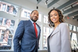 Alt text: A man and a woman in business attire smiling confidently inside an office with large windows.