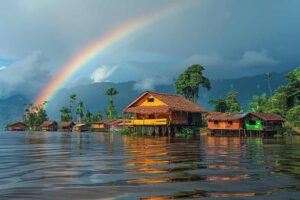 Alt text: A row of stilt houses on a calm body of water with a vibrant rainbow arcing across the sky.