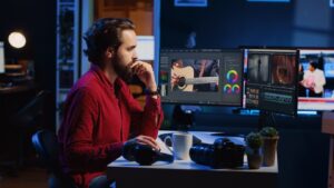 Alt text: A man in a red shirt is deeply focused on editing video content on dual monitors at his desk, which is equipped with headphones, a camera, and a coffee mug.