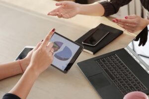 Alt text: Two people discussing a pie chart displayed on a tablet, with a laptop and notebook on the table.